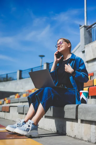 Modern Park Caddesi 'nde dizüstü bilgisayarı olan genç ve güzel bir kadın uzaktan kumandalı akıllı telefon kullanıyor. Yaz tarzı gülümseme gözlüğü takıyor.