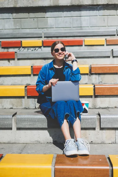 Modern Park Caddesi 'nde dizüstü bilgisayarı olan genç ve güzel bir kadın uzaktan kumandalı akıllı telefon kullanıyor. Yaz tarzı gülümseme gözlüğü takıyor.