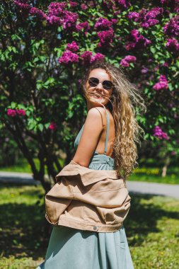 Yazlık elbiseli güzel bir kıvırcık kadın koşar ve sevinir, güneş gözlüğü takar. Mor bir leylak çalısının arka planında