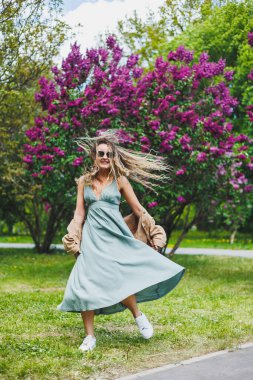 Yazlık elbiseli güzel bir kıvırcık kadın koşar ve sevinir, güneş gözlüğü takar. Mor bir leylak çalısının arka planında