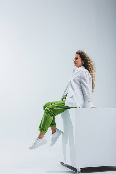 Позитивная женщина с вьющимися волосами в белой светлой хлопчатобумажной рубашке и брюках на белом изолированном фоне. Женская повседневная одежда.