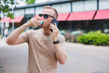 Sakallı, gülümseyen, beyaz gömlekli ve güneş gözlüklü şık bir adam şehrin sokaklarında duruyor ve cep telefonundan arıyor..