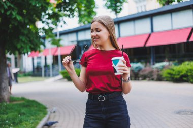 Sırt çantalı, gülümseyen ve bakışlarını kaçıran genç bir bayan. Şehir caddesinde yürürken ve taze kahvenin tadını çıkarırken.