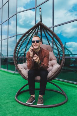 İşten sonra ofis binasının terasında oturan, tişörtlü neşeli genç bir adam, kablosuz internet kullanıyor, gözlüklü pozitif bir erkek telefonda konuşuyor.