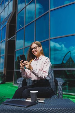 Klasik gözlüklü ve modaya uygun giyinmiş güzel bir kadın cep telefonundan mesaj okuyor, modern cam bir binanın yanında duruyor..