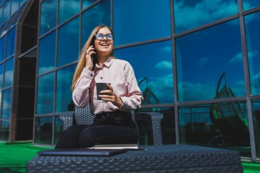 Klasik gözlüklü ve modaya uygun giyinmiş güzel bir kadın cep telefonundan mesaj okuyor, modern cam bir binanın yanında duruyor..