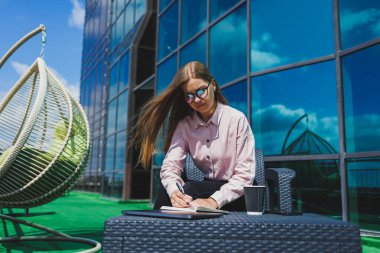 Not defteriyle sokak kafesinde poz veren neşeli bayan öğrenci ders kitabı ve kahve bardağıyla dışarıda gülümseyen başarılı kadın.