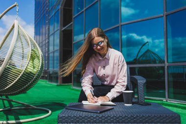Not defteriyle sokak kafesinde poz veren neşeli bayan öğrenci ders kitabı ve kahve bardağıyla dışarıda gülümseyen başarılı kadın.