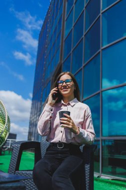Klasik gözlüklü ve modaya uygun giyinmiş güzel bir kadın cep telefonundan mesaj okuyor, modern cam bir binanın yanında duruyor..