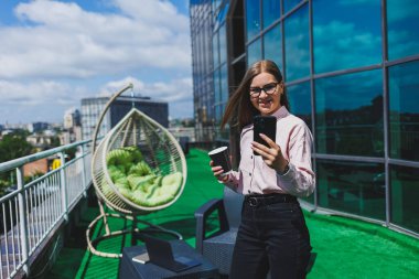 Ofis balkonunda kahve molası sırasında akıllı telefondan SMS okuyan gülümseyen iş kadınının portresi. Rahatlamak için modern teras ofiste.