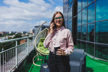 Ofis balkonunda kahve molası sırasında akıllı telefondan SMS okuyan gülümseyen iş kadınının portresi. Rahatlamak için modern teras ofiste.