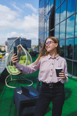 Ofis balkonunda kahve molası sırasında akıllı telefondan SMS okuyan gülümseyen iş kadınının portresi. Rahatlamak için modern teras ofiste.