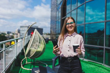 Ofis balkonunda kahve molası sırasında akıllı telefondan SMS okuyan gülümseyen iş kadınının portresi. Rahatlamak için modern teras ofiste.