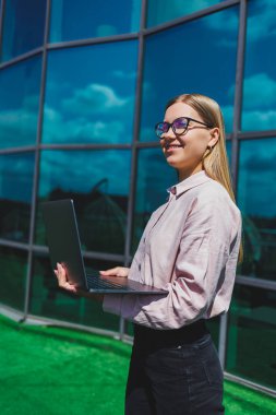 Sıradan kıyafetler ve moda gözlüklerle konsantre olmuş neşeli kadın elinde dizüstü bilgisayarla gökdelenlerin önünde duruyor, gülümsüyor ve dizüstü bilgisayarla internette geziniyor.