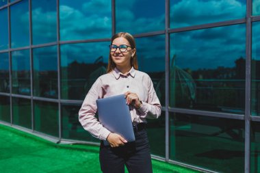 Sıradan kıyafetler ve moda gözlüklerle konsantre olmuş neşeli kadın elinde dizüstü bilgisayarla gökdelenlerin önünde duruyor, gülümsüyor ve dizüstü bilgisayarla internette geziniyor.