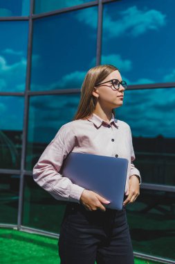 Sıradan kıyafetler ve moda gözlüklerle konsantre olmuş neşeli kadın elinde dizüstü bilgisayarla gökdelenlerin önünde duruyor, gülümsüyor ve dizüstü bilgisayarla internette geziniyor.
