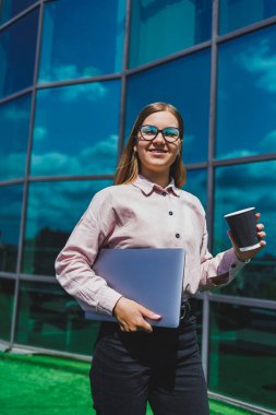Elinde kahve olan bardaklı bir kadın, gökdelene karşı. Modern bina, finans merkezi, şehir manzarası. Gözlüklü başarılı bir bayan girişimci.