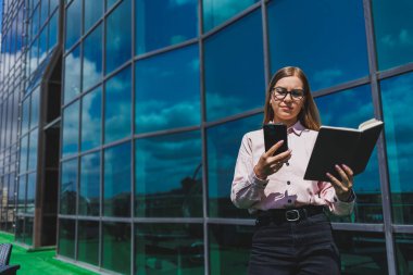 Hoş, neşeli bir kadın yönetici günlük kıyafetler ve şık gözlükler içinde elinde bir defterle bir gökdelenin arka planında duruyor, gülümsüyor ve telefonda konuşuyor.