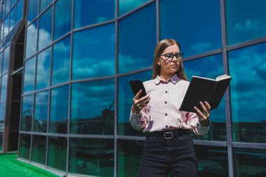 Hoş, neşeli bir kadın yönetici günlük kıyafetler ve şık gözlükler içinde elinde bir defterle bir gökdelenin arka planında duruyor, gülümsüyor ve telefonda konuşuyor.