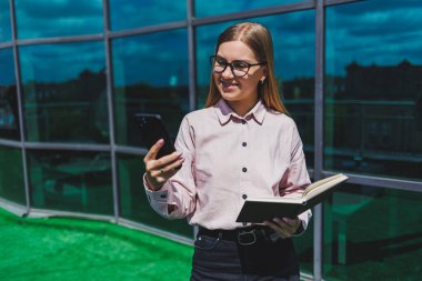 Hoş, neşeli bir kadın yönetici günlük kıyafetler ve şık gözlükler içinde elinde bir defterle bir gökdelenin arka planında duruyor, gülümsüyor ve telefonda konuşuyor.