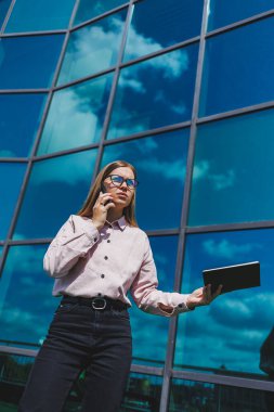 Hoş, neşeli bir kadın yönetici günlük kıyafetler ve şık gözlükler içinde elinde bir defterle bir gökdelenin arka planında duruyor, gülümsüyor ve telefonda konuşuyor.