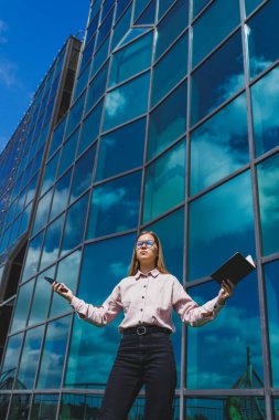 Hoş, neşeli bir kadın yönetici günlük kıyafetler ve şık gözlükler içinde elinde bir defterle bir gökdelenin arka planında duruyor, gülümsüyor ve telefonda konuşuyor.
