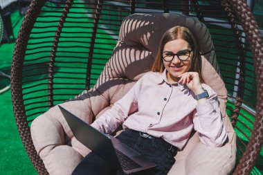 Çekici bir kadın elinde dizüstü bilgisayarla balkonda oturur ve dikkatle ekrana bakar. Serbest giyinmiş güzel bir kız balkonda bir sandalyede otururken dizüstü bilgisayarda çalışıyor..