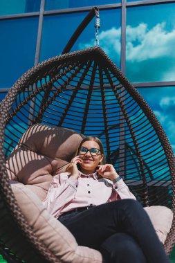 Güzel, neşeli bir bayan yönetici, günlük kıyafetleri ve modaya uygun gözlükleriyle ofis terasında oturuyor ve dinleniyor. Çalışma gününden sonra rahatla.