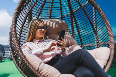 Güzel, neşeli bir bayan yönetici, günlük kıyafetleri ve modaya uygun gözlükleriyle ofis terasında oturuyor ve dinleniyor. Çalışma gününden sonra rahatla.