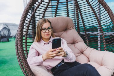Klasik gözlüklü ve modaya uygun giyinmiş güzel bir kadın cep telefonundan mesaj okuyor, modern cam bir binanın yanında duruyor..