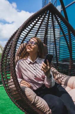 Güzel, neşeli bir bayan yönetici, günlük kıyafetleri ve modaya uygun gözlükleriyle ofis terasında oturuyor ve dinleniyor. Çalışma gününden sonra rahatla.
