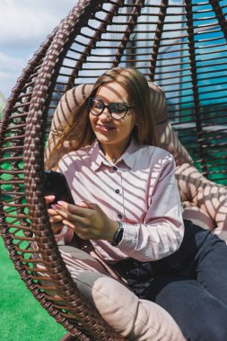 Güzel, neşeli bir bayan yönetici, günlük kıyafetleri ve modaya uygun gözlükleriyle ofis terasında oturuyor ve dinleniyor. Çalışma gününden sonra rahatla.
