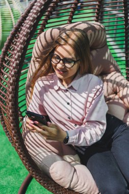 Güzel, neşeli bir bayan yönetici, günlük kıyafetleri ve modaya uygun gözlükleriyle ofis terasında oturuyor ve dinleniyor. Çalışma gününden sonra rahatla.