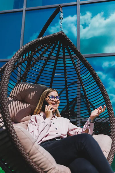 Güzel, neşeli bir bayan yönetici, günlük kıyafetleri ve modaya uygun gözlükleriyle ofis terasında oturuyor ve dinleniyor. Çalışma gününden sonra rahatla.