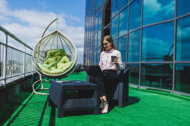İş akışı için dizüstü bilgisayarı olan neşeli bir serbest çalışan sokak kafesinde poz veren, bilgisayar ve kahve bardağıyla gülümseyen başarılı bir kadın.
