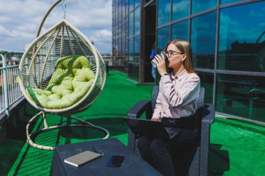 İş akışı için dizüstü bilgisayarı olan neşeli bir serbest çalışan sokak kafesinde poz veren, bilgisayar ve kahve bardağıyla gülümseyen başarılı bir kadın.