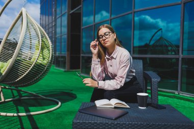 Not defteriyle sokak kafesinde poz veren neşeli bayan öğrenci ders kitabı ve kahve bardağıyla dışarıda gülümseyen başarılı kadın.