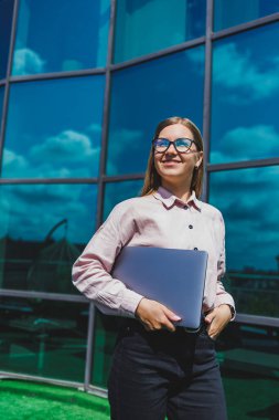 Sıradan kıyafetler ve moda gözlüklerle konsantre olmuş neşeli kadın elinde dizüstü bilgisayarla gökdelenlerin önünde duruyor, gülümsüyor ve dizüstü bilgisayarla internette geziniyor.