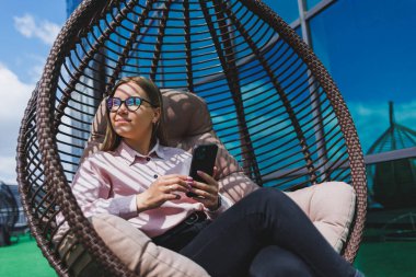 Güzel, neşeli bir bayan yönetici, günlük kıyafetleri ve modaya uygun gözlükleriyle ofis terasında oturuyor ve dinleniyor. Çalışma gününden sonra rahatla.