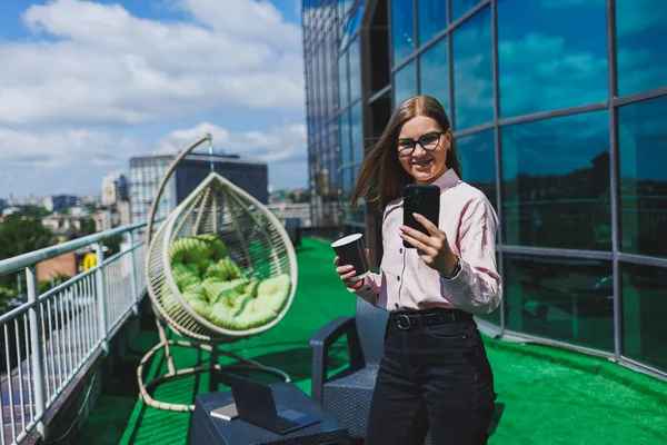 Ofis balkonunda kahve molası sırasında akıllı telefondan SMS okuyan gülümseyen iş kadınının portresi. Rahatlamak için modern teras ofiste.