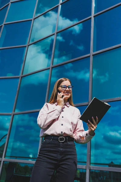 A nice cheerful woman manager in casual clothes and fashionable glasses stands against the backdrop of a skyscraper with a notebook in her hands, smiles and talks on the phone