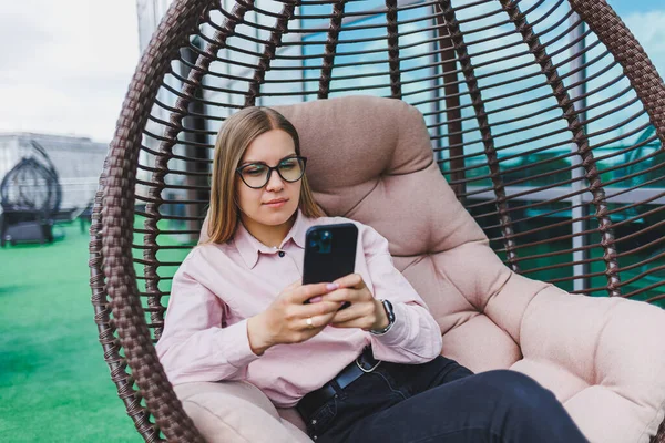 Klasik gözlüklü ve modaya uygun giyinmiş güzel bir kadın cep telefonundan mesaj okuyor, modern cam bir binanın yanında duruyor..