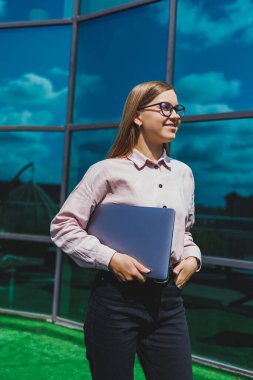 Sıradan kıyafetler ve moda gözlüklerle konsantre olmuş neşeli kadın elinde dizüstü bilgisayarla gökdelenlerin önünde duruyor, gülümsüyor ve dizüstü bilgisayarla internette geziniyor.