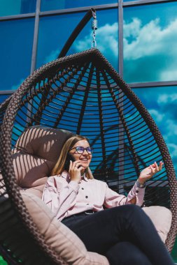 Güzel, neşeli bir bayan yönetici, günlük kıyafetleri ve modaya uygun gözlükleriyle ofis terasında oturuyor ve dinleniyor. Çalışma gününden sonra rahatla.