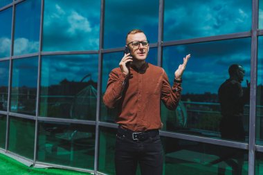 Görme düzeltmesi için klasik gözlüklü mutlu adam cep telefonuyla terasta konuşuyor, gülen blogcu sosyal medya sitesine göz atıyor.