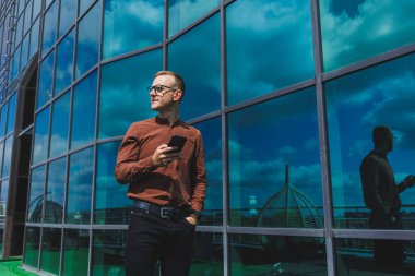 Görme düzeltmesi için klasik gözlüklü mutlu adam cep telefonuyla terasta konuşuyor, gülen blogcu sosyal medya sitesine göz atıyor.