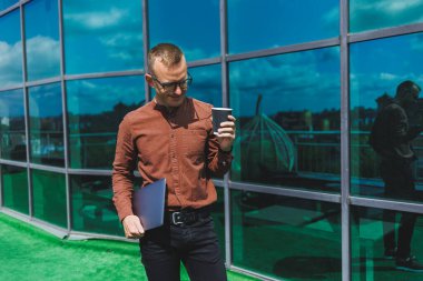 Gözlüklü, dizüstü bilgisayarı ve bir bardak kahvesi olan yakışıklı bir adam bir ofisin terasında duruyor. genç akıllı geliştirici