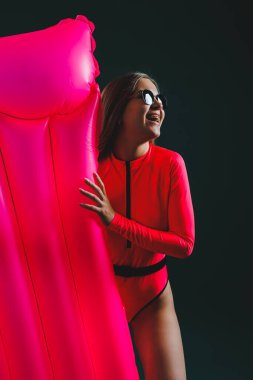 Pembe mayo ve güneş gözlüğü takmış güzel bir kadın yeşil arka planda pembe şişme yatakla poz veriyor.