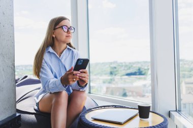 Klasik gözlüklü serbest çalışan bir kadın modern bir çalışma alanında otururken telefonla konuşuyor ve gülümsüyor. Gözlüklü bir kadın, iletişim için boş vakitlerin keyfini çıkarıyor.