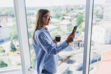 Modern kadın, büyük bir pencerede durup kahve içerken telefonla konuşuyor. Modern başarılı kadın kavramı. Açık bir ofiste genç bir kız